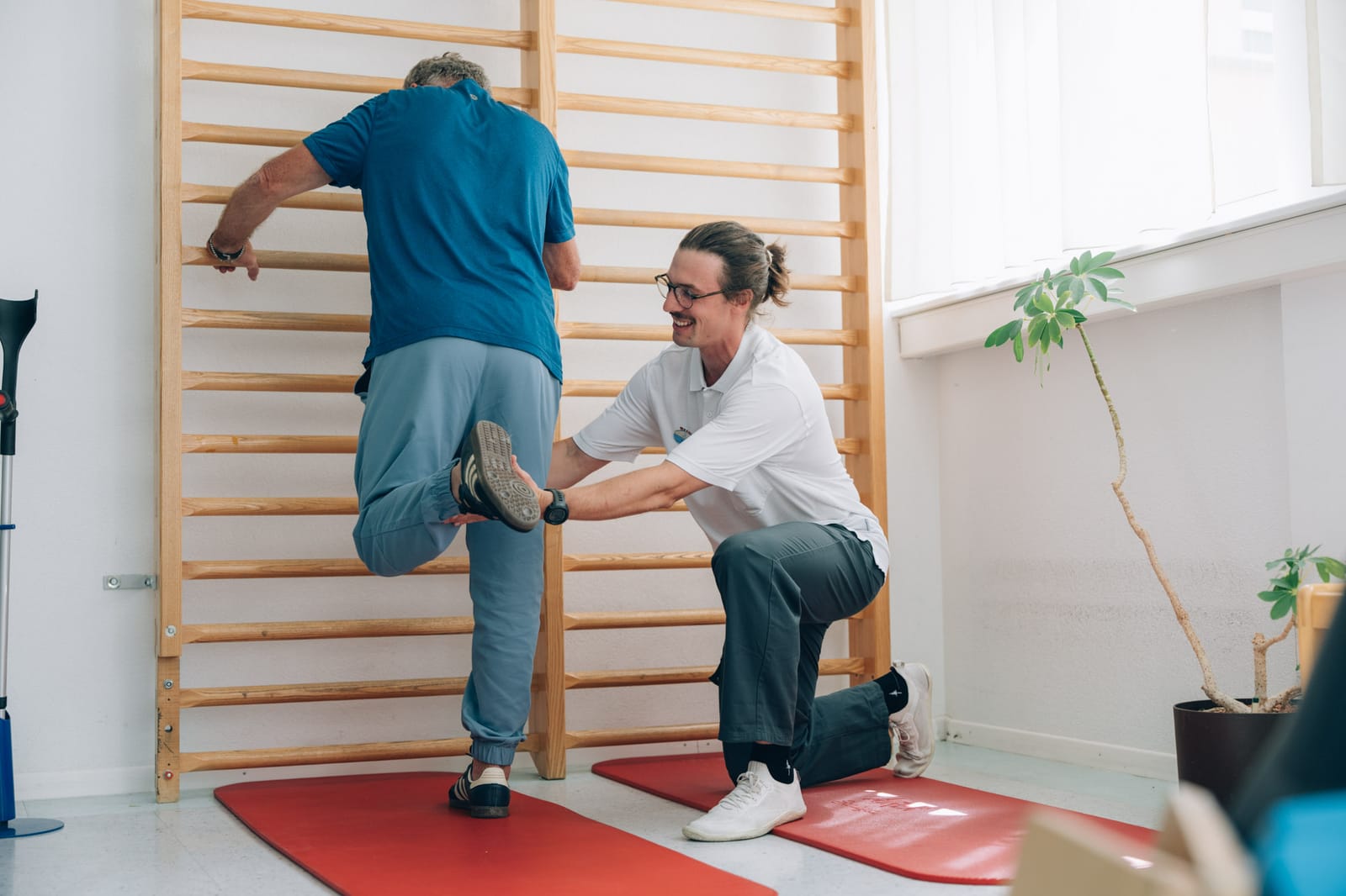Ein Patient hält sich von hinten gesehen an der Sprossenwand und hebt ein Bein; ein Therapeut kniet daneben und führt den Fuss