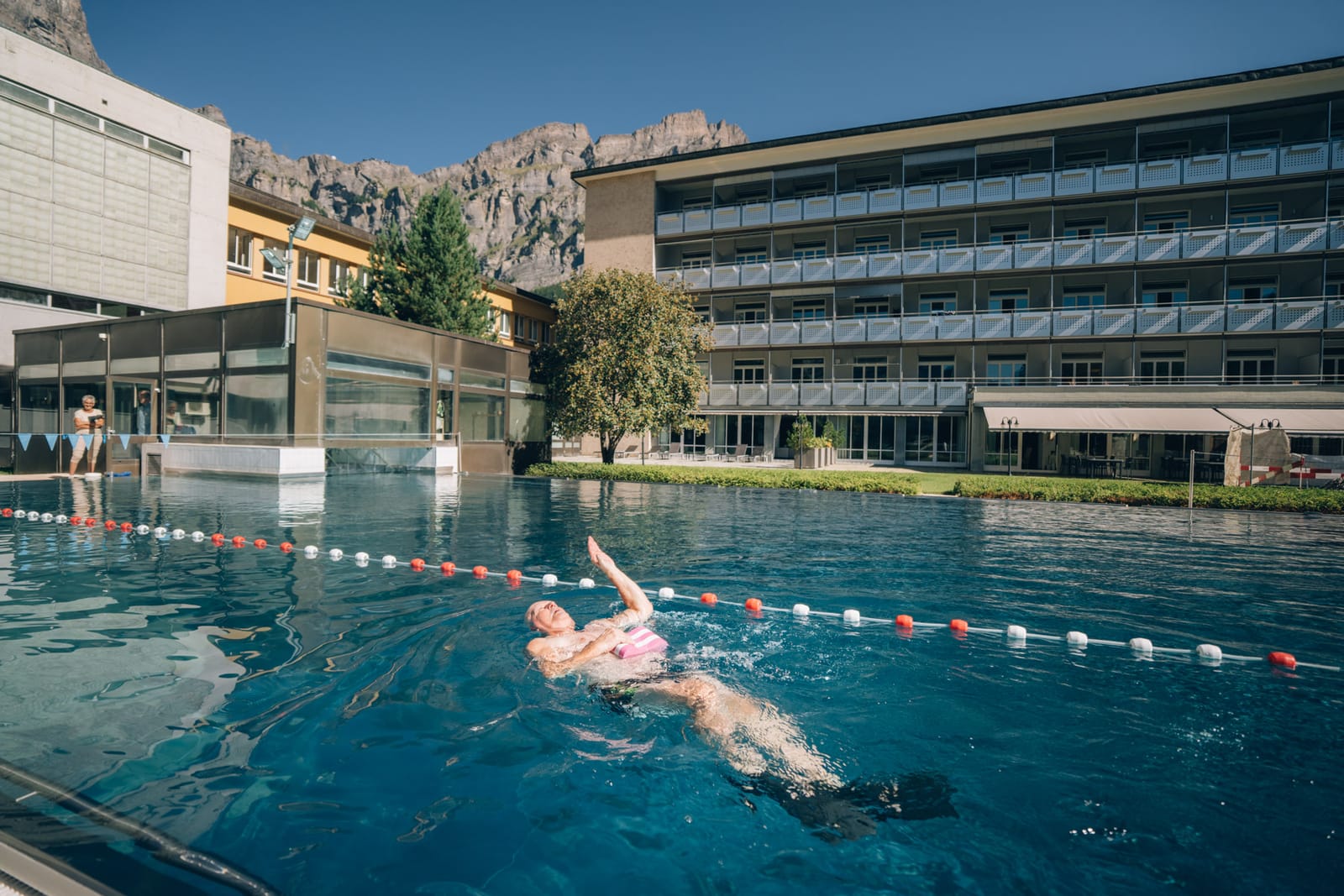 Eine Person schwimmt auf dem Rücken im Aussenbecken, dahinter die Gebäude der Klinik und die Felsen